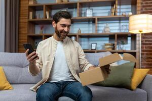 een jong Mens is zittend Bij huis Aan de bank, Holding een telefoon in zijn hand, gelukkig naar hebben ontvangen een pakket door levering, opening de doos. foto