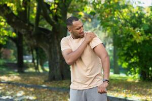 een Mens gewond zijn schouder gedurende een geschiktheid klas, een Afro-Amerikaans Mens gewond zichzelf terwijl jogging in de park, strekt zich uit zijn arm en massages zijn zeer spieren. foto