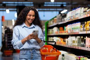 een jong Latijns Amerikaans vrouw is boodschappen doen in een supermarkt tussen de schappen met goederen, een vrouw toepassingen een toepassing Aan haar telefoon naar vergelijken prijzen. foto