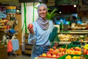 portret van een vrouw koper in een supermarkt, een moslim vrouw in een hijab is glimlachen gelukkig en op zoek Bij de camera, kiezen appels en fruit in een groot kruidenier op te slaan. foto