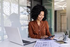 geslaagd gelukkig accountant financier met een rekenmachine bereidt zich voor een financieel verslag doen van en vult in tafel vormen, zakenvrouw werken met papieren, contracten binnen kantoor, vrouw arbeider met een laptop. foto