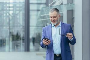 geslaagd en gelukkig senior grijs haar zakenman investeerder wandelen buiten kantoor gebouw, Mens Holding telefoon, bankier gebruik makend van smartphone, typen bericht en lezing nieuws foto