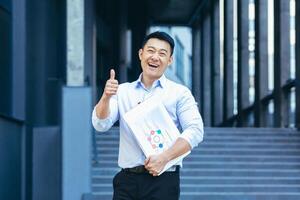 geslaagd Aziatisch zakenman buiten kantoor lachend, duimen omhoog bevestigend foto