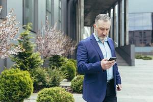 nadenkend senior en ervaren Mens buiten kantoor lezing nieuws Aan telefoon, zakenman buiten kantoor gebruik makend van smartphone terwijl wandelen in de buurt gebouw foto