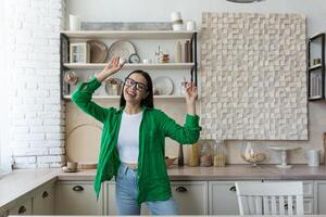 gelukkig jong mooi vrouw in bril en groen overhemd dansen en het zingen Bij huis in de keuken. resting van huiswerk, ontspannen, genieten van tijd alleen foto