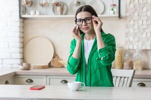 depressie, paniek aandoeningen. een jong mooi vrouw in bril en een groen overhemd is huilen Bij huis, zittend Bij de keuken tafel, afvegen haar tranen met een servet. hij voelt slechte, voelt angst en apathie foto