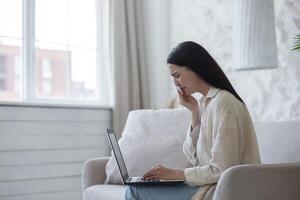 quarantaine. oorlog. een mooi jong vrouw zit Bij huis Aan de bankstel met een laptop, praat online via koppeling met haar familie en vrienden, huilt, mist hen. doekjes tranen met een servet. foto