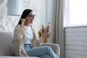 gelukkig jong mooi vrouw in bril zittend Aan sofa Bij huis met telefoon in handen. viert een geslaagd zege en winnend de loterij, schreeuwt wauw, points met zijn hand- ja. foto