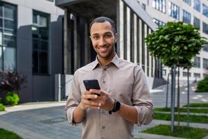 portret van jong zakenman, Mens glimlachen op zoek Bij camera Holding telefoon, arbeider gebruik makend van app Aan smartphone terwijl wandelen in stad dichtbij omhoog buiten kantoor gebouw. foto