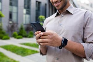 dichtbij omhoog van man's handen, zakenman in overhemd Holding telefoon, gebruik makend van app Aan smartphone, wandelen in stad buiten kantoor gebouw. foto