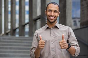 zelfverzekerd jong Mens in een gewoontjes overhemd geven duimen omhoog met een glimlach, buitenshuis stappen in de achtergrond, uitdrukken succes en positiviteit. foto