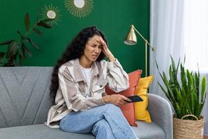 een jong vrouw is getoond zittend Aan een grijs bankstel in een huis leven kamer, drukken haar vingers naar haar tempels, weergeven tekens van een hoofdpijn of spanning terwijl Holding een afgelegen controle. foto