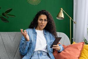 verward vrouw in bril Holding telefoon, gebaren onzekerheid, zittend Aan bankstel Bij huis met elegant decor. foto