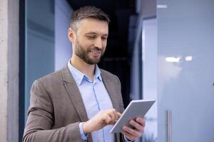 volwassen ervaren zakenman binnen kantoor glimlachen tevreden, senior Mens Holding tablet computer in handen, baas in bedrijf pak gebruik makend van analytisch financieel sollicitatie. foto