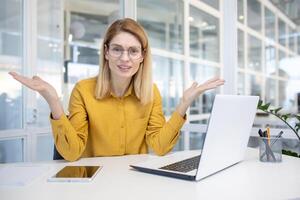 een verward vrouw vervelend bril en een geel overhemd werken Aan haar laptop in een modern kantoor, uitdrukken verrassing of onzekerheid. foto