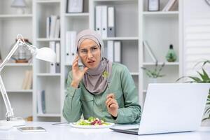 uitgeput moslim vrouw in hijab zittend Bij bureau en aan het eten salade terwijl werken Aan laptop. moe vrouw ervaren hoofdpijn in kantoor met wit schappen en decoratief artikelen. foto