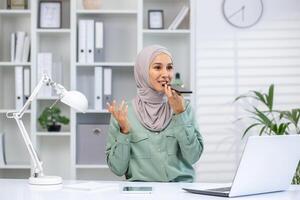 een vrolijk moslim vrouw vervelend een hijab spreekt in haar telefoon, opname een audio bericht Bij haar goed georganiseerd kantoor ruimte versierd met planten en een hedendaags wit lamp. foto