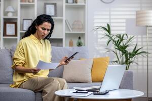 ontevreden en teleurgesteld vrouw achter papier werk zittend Aan sofa Bij huis, spaans vrouw Holding nut rekeningen, schuldeiser cheques, betalen van streek gebruik makend van telefoon app. foto