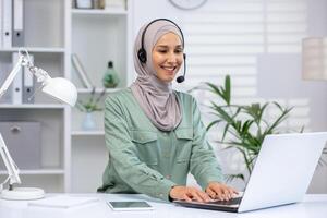 een vrolijk professioneel moslim vrouw in een hijab verloofd in een klant onderhoud telefoongesprek, gebruik makend van een koptelefoon en laptop in een modern kantoor. foto