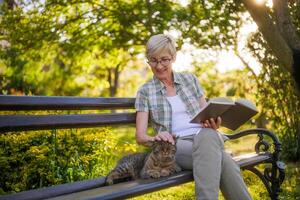 gelukkig senior vrouw geniet lezing boek en uitgeven tijd met haar kat terwijl zittend Aan een bank in haar tuin. foto