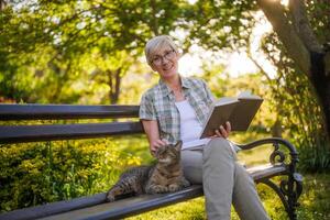 gelukkig senior vrouw geniet lezing boek en uitgeven tijd met haar kat terwijl zittend Aan een bank in haar tuin foto