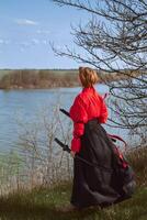 een vrouw met kort rood haar- in een zwart en rood Hanfu en twee katana's looks in de afstand tegen de achtergrond van een voorjaar rivier- foto