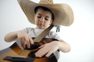 jongen 7 jaren oud in een reusachtig cowboy hoed Toneelstukken de gitaar detailopname Aan een wit achtergrond genoegen aan het leren naar Speel musical instrumenten muziek- school- festivals tonen programma voor kinderen foto