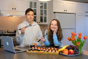 hebben pret tiener- jongen en meisje aan het kijken film Aan laptops en aan het eten sushi in keuken voedsel levering pret tijdverdrijf aangenaam verhouding eerste datum vrienden klasgenoten samen leren naar koken Aziatisch voedsel foto