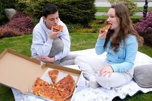 jongen en meisje tieners eten pizza zittend Aan een deken in natuur sportkleding adolescentie communicatie vrienden eerste datum heerlijk voedsel snel voedsel pizza levering pizza meenemen park foto