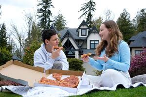 tiener jongen en meisje pratend aan het eten pizza zittend in natuur tiener wandelen ontspannende in vers lucht eerste stralen van zon rust uit in voorjaar Gaan uit zitten Aan gras communiceren offline Bij huis terug voorjaar foto