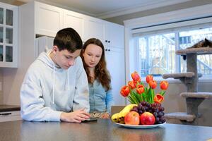 jongens meisje in de keuken op zoek voor voedsel levering in telefoon stil leven broer en zus zonder ouders eten snel voedsel levering Aan de tafel fruit bloemen kat resting Aan boom kat wit en meubilair foto