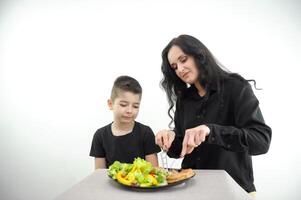 mam geeft les zoon naar eten Rechtsaf snel voedsel voor kinderen is slecht voor Gezondheid mam bezuinigingen uit stuk van vlees Aan bord salade vers alleen maar gekookt voedsel lunch avondeten Aan wit achtergrond zwart kleren nodig hebben naar eten Rechtsaf foto