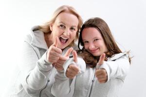 dichtbij omhoog foto mooi twee mensen bruin haren mam weinig dochter tonen duimen in lucht aanbevelen knuffelen het beste vrienden kijken vergelijkbaar vervelend wit t-shirts geïsoleerd helder blauw achtergrond