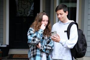 tieners tonen interessant nieuws jongen en meisje met aktentas telefoon Bij deur Bij huis glimlachen paar op zoek in smartphone samen. mooi liefhebbend paar lachend en aan het kijken iets Aan mobiel telefoon foto