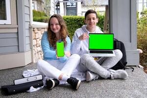 tiener jongen en tiener- meisje zittend buitenshuis met mappen rugzak Holding laptop en telefoon groen scherm chroma sleutel advertentie kijken hier school- presentatie Aan computer kind klaar voor les. tiener laptop. foto