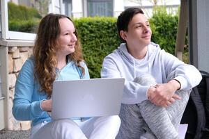 tieners zitten Aan veranda na school- kijken in een richting in handen met laptop eerste liefde, jong tieners geliefden paar aan het studeren laptop, jongen en meisje aan het doen huiswerk, eerste daten, bloem en kinderen liefde. foto