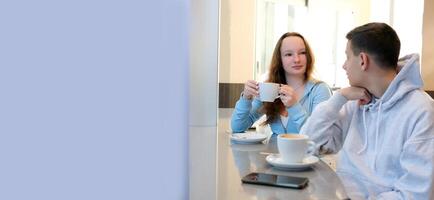 jong mensen jongen meisje zittend in een cafe tiener chatten drinken koffie wit kop in hand- lachend hebben pret uitgeven tijd samen leerling kantine restaurant cafe school- zitten door venster wit kader foto