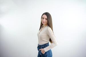 slank schattig jong meisje in jeans Aan een wit achtergrond in de studio aantrekkelijk brunette zetten haar handen in haar terug zakken, schattig poseren tegen een beige muur achtergrond foto