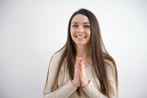 vouwen uw handen in voorkant van uw borst net zo in gebed zorgeloos spaans vrouw glimlachen en op zoek Bij camera staand Aan grijs achtergrond. mooi multi-etnisch meisje lachend. foto