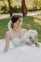 een vrouw in een wit jurk is zittend Aan de gras Holding een boeket van bloemen foto