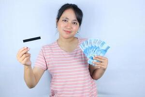 portret van een glimlachen Aziatisch jong vrouw Holding contant geld roepia en credit kaart Aan wit achtergrond foto