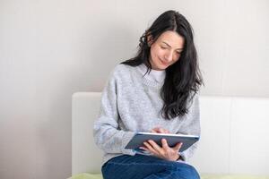 glimlachen midden oud vrouw in grijs trui houdt een tablet, lezing of werken, illustreren digitaal geletterdheid en comfort Bij middelbare leeftijd, net zo goed net zo midlife en haar uitdagingen. foto