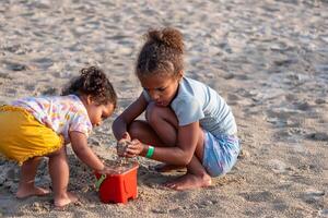 twee jong Afrikaanse Amerikaans kinderen, geabsorbeerd in vulling een emmer met zand, delen een moment van coöperatie Speel Aan de strand, broer of zus liefde en nationaal broers en zussen dag. foto