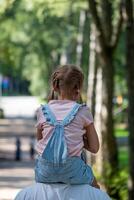 een jong meisje is krijgen gedragen Aan haar vaders schouders geniet een zonnig dag in een met bomen omzoomd park, een momentopname van onschuld en de gemakkelijk vreugden van jeugd. foto