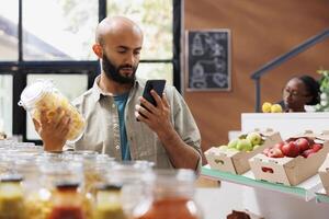 gebaard Mens in detailopname gebruik makend van smartphone naar zoeken voor recepten en prijzen Bij dichtbij eco vriendelijk winkel. midden- oostelijk klant browsen Aan internet voor advies Aan duurzame gezond leven. foto