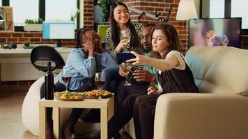 multi-etnisch vrienden nemen groep afbeeldingen met mobiele telefoon Bij appartement bijeenkomst. gelukkig mensen in leven kamer poseren voor foto, maken grappig gezichten en drinken alcoholisch dranken foto