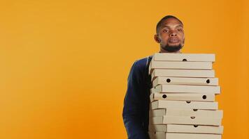 onhandig postbode dropping een reusachtig stack van pizza dozen in studio, verspilling een veel van voedsel en maken een troep. jong verontrust Mens maakt een vergissing en morsen een groot stapel van snel voedsel. camera b. foto