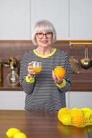 vrolijke, mooie senior lachende vrouw in gestreepte trui die sinaasappelsap drinkt terwijl ze in de keuken staat. gezonde, sappige levensstijl, thuis, senior mensen concept.