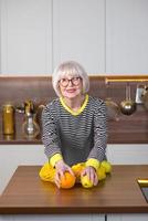 vrolijke mooie senior lachende vrouw in gestreepte trui die citroenen vasthoudt voor limonade terwijl ze in de keuken staat. gezonde, sappige levensstijl, thuis, senior mensen concept. foto