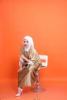 vol lengte studio portret van volwassen elegant moslim Aziatisch vrouw krom lichaam vervelend kaftan hijaab, hoog hakken zittend Aan een stoel geïsoleerd Aan oranje achtergrond. mode, levensstijl, reclame concept foto